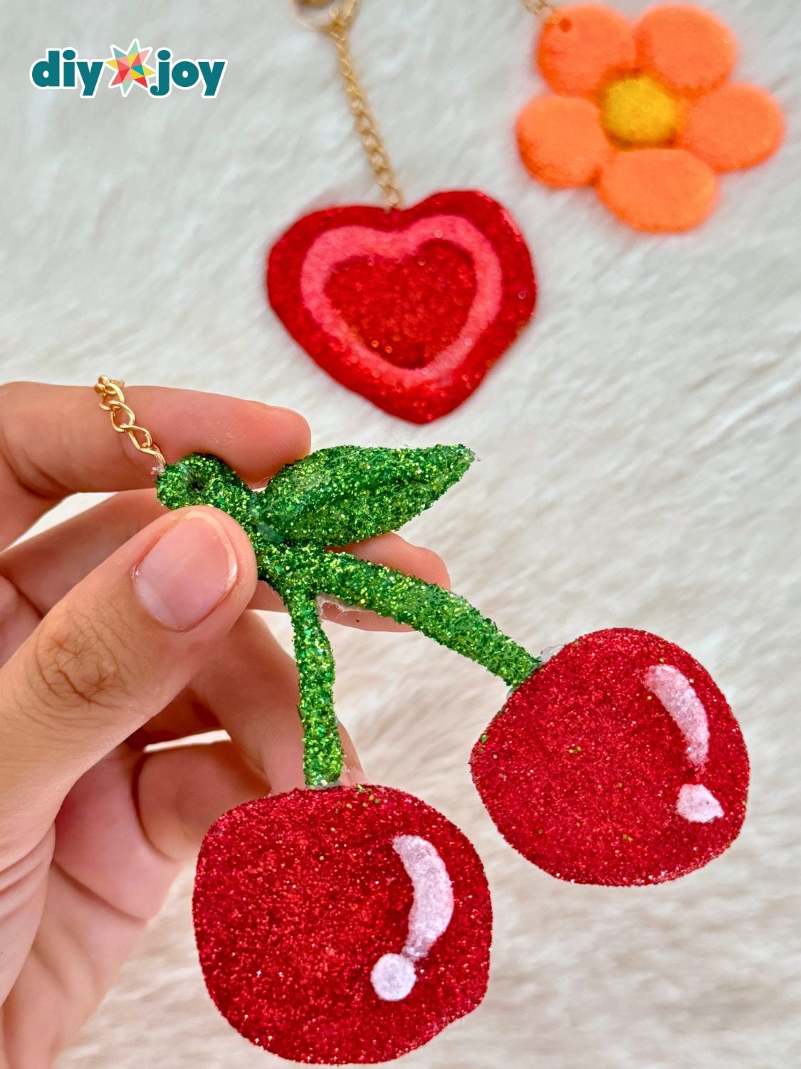 Hand holding a red glitter cherry keychain on a gold chain with a glitter heart and orange daisy keychain on white fur