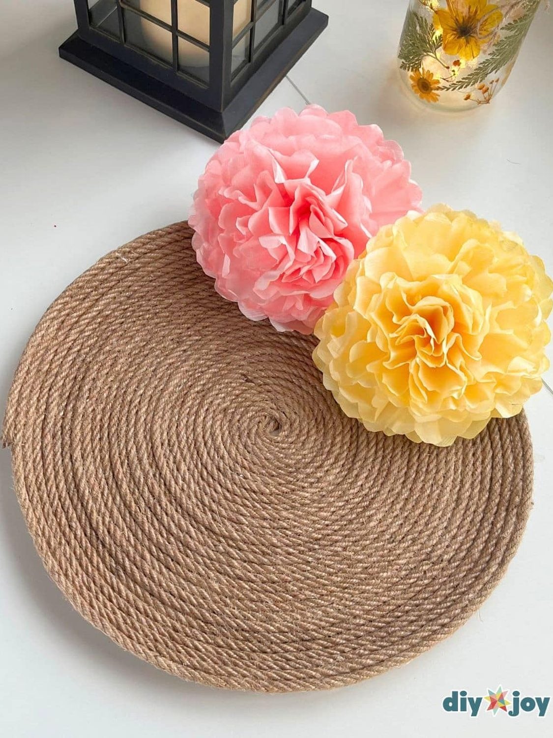 Round woven jute rope tray on a white table holding a pink and a yellow paper flower beside a black lantern