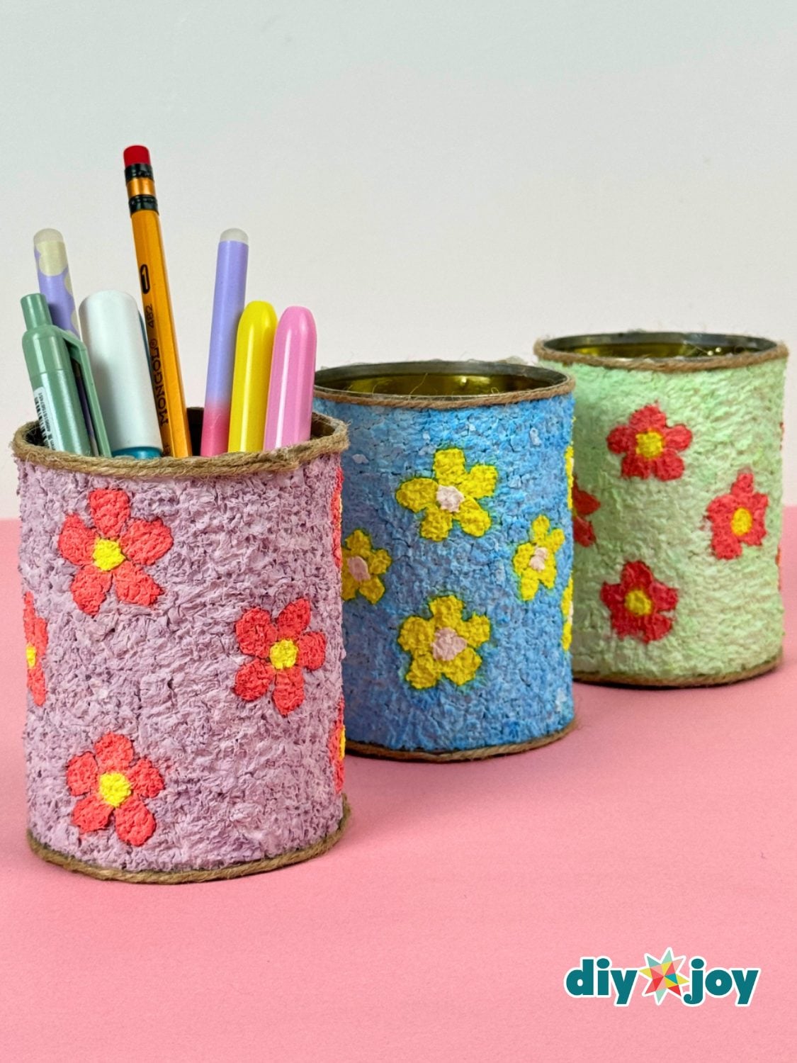 Three tin cans covered in pastel tissue paper pulp with painted flowers and twine rims, holding pens on a pink table