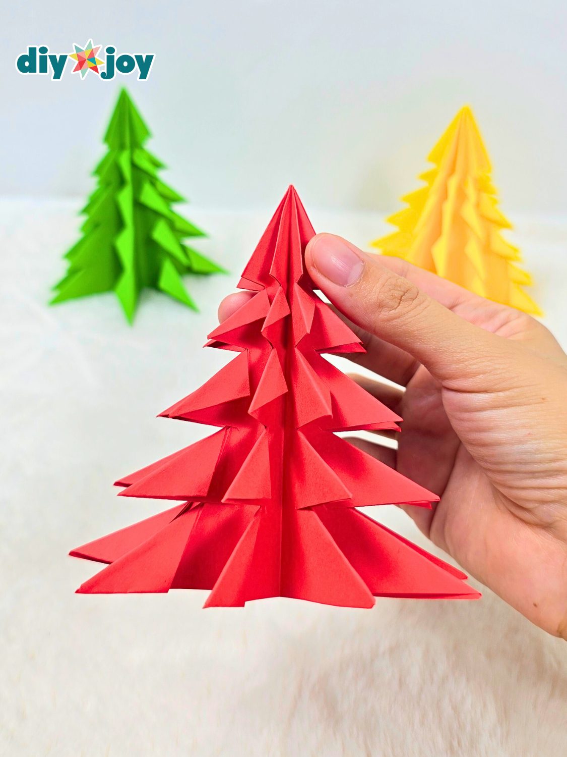 A hand holding a green three dimensional origami paper Christmas tree with layered pointed tiers on a plain background