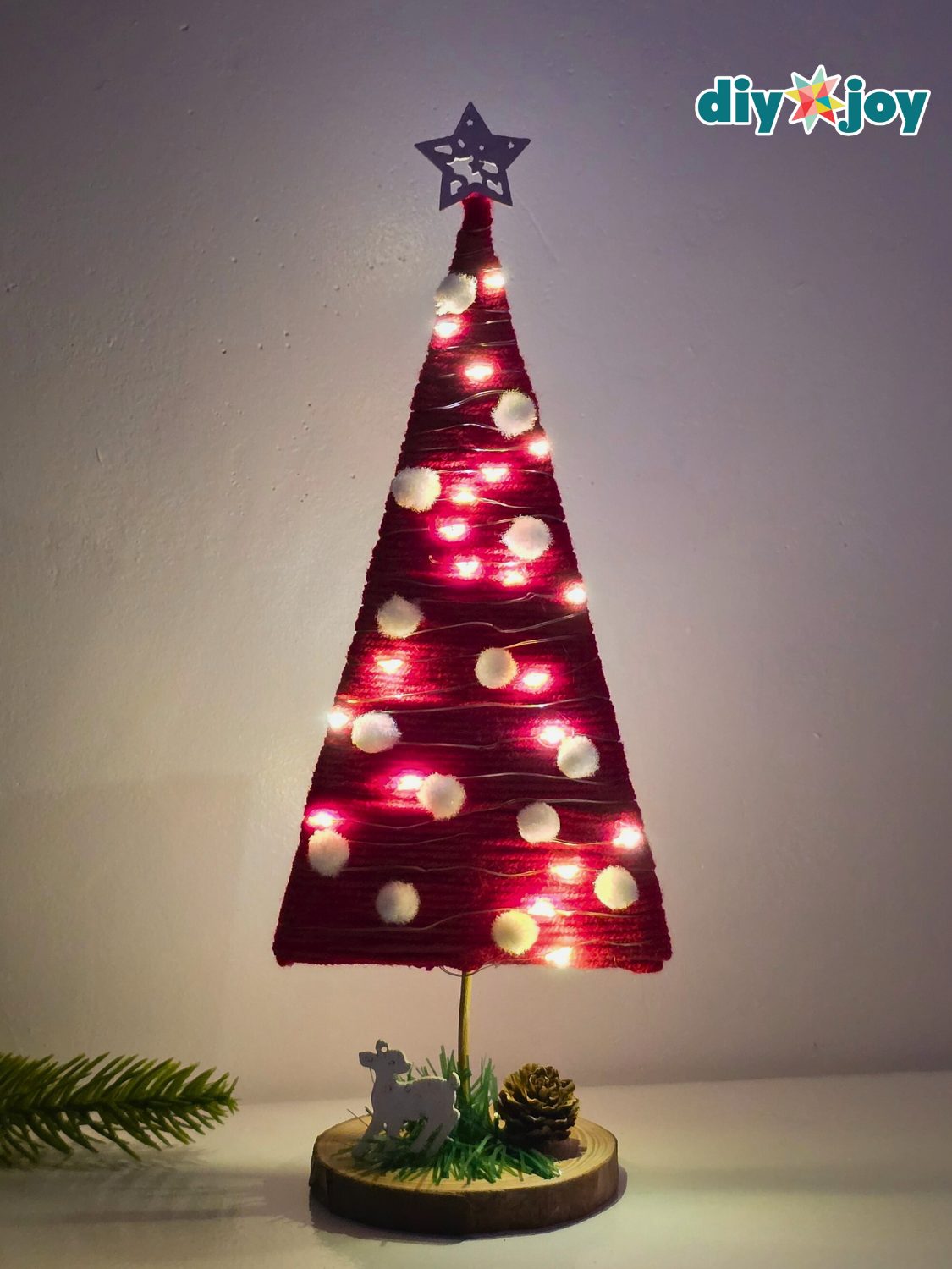 A mini red yarn wrapped Christmas tree with white pom poms and glowing fairy lights on a wood slice base with a tiny deer