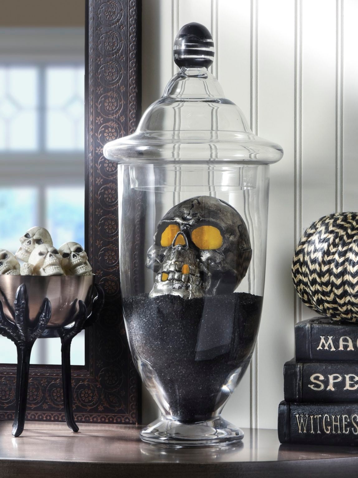 Black skull glowing inside a clear apothecary jar over black granules, with small skulls and spell books nearby