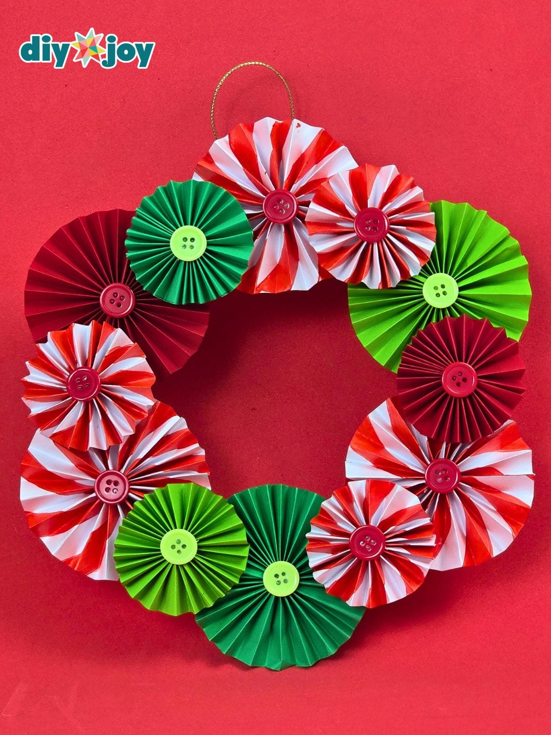 Wreath made of red, green and peppermint striped paper rosettes with button centers hanging from a gold cord on a red wall