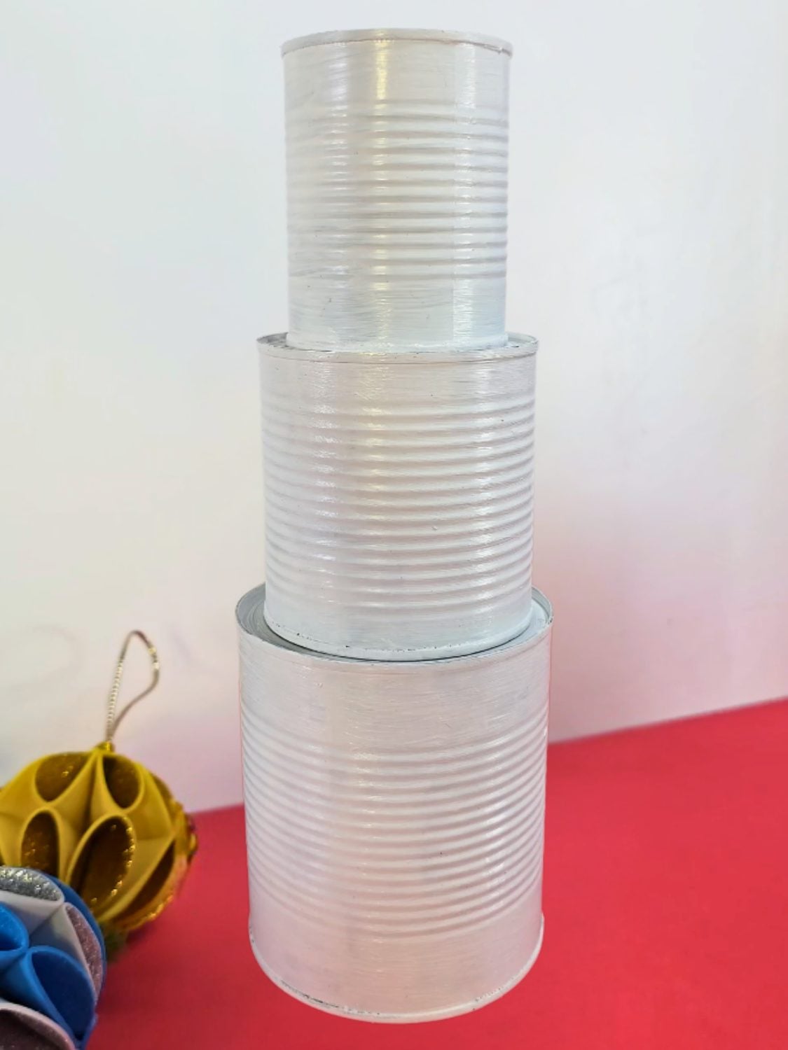 Three tin cans painted white and stacked into a snowman shape on a red surface beside paper ornaments