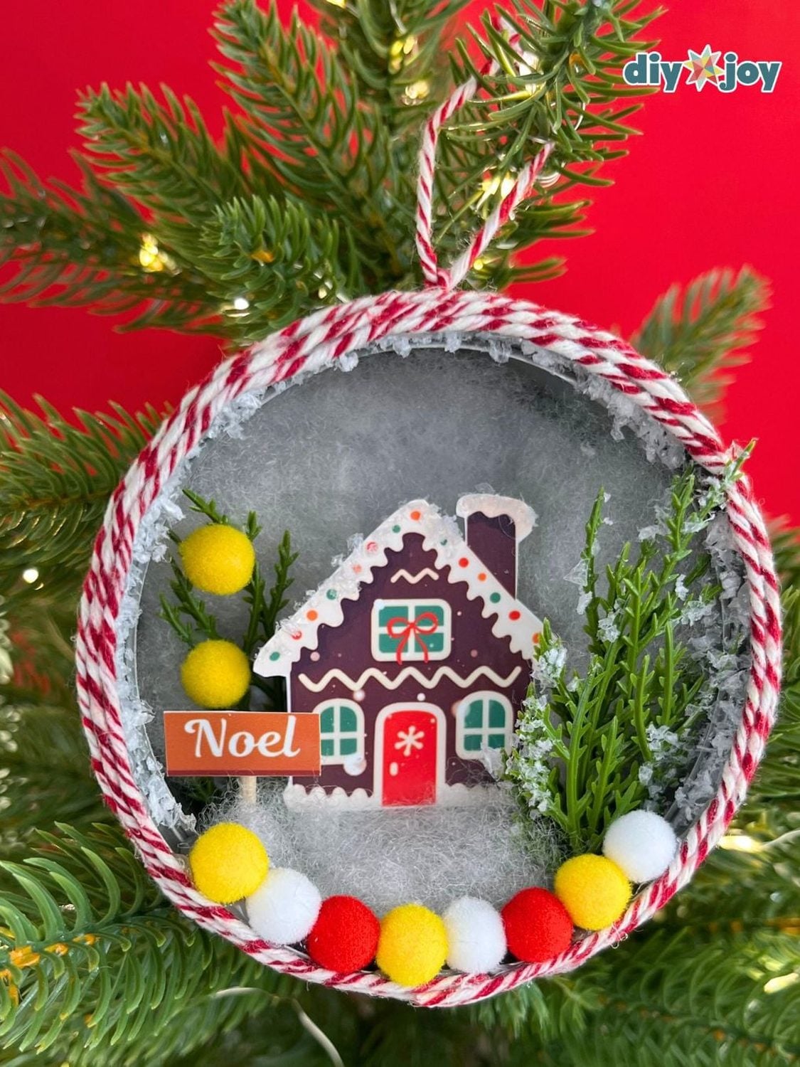 Tuna can ornament with a gingerbread house scene, faux pine, pom poms and red and white twine on the rim, hung on a tree
