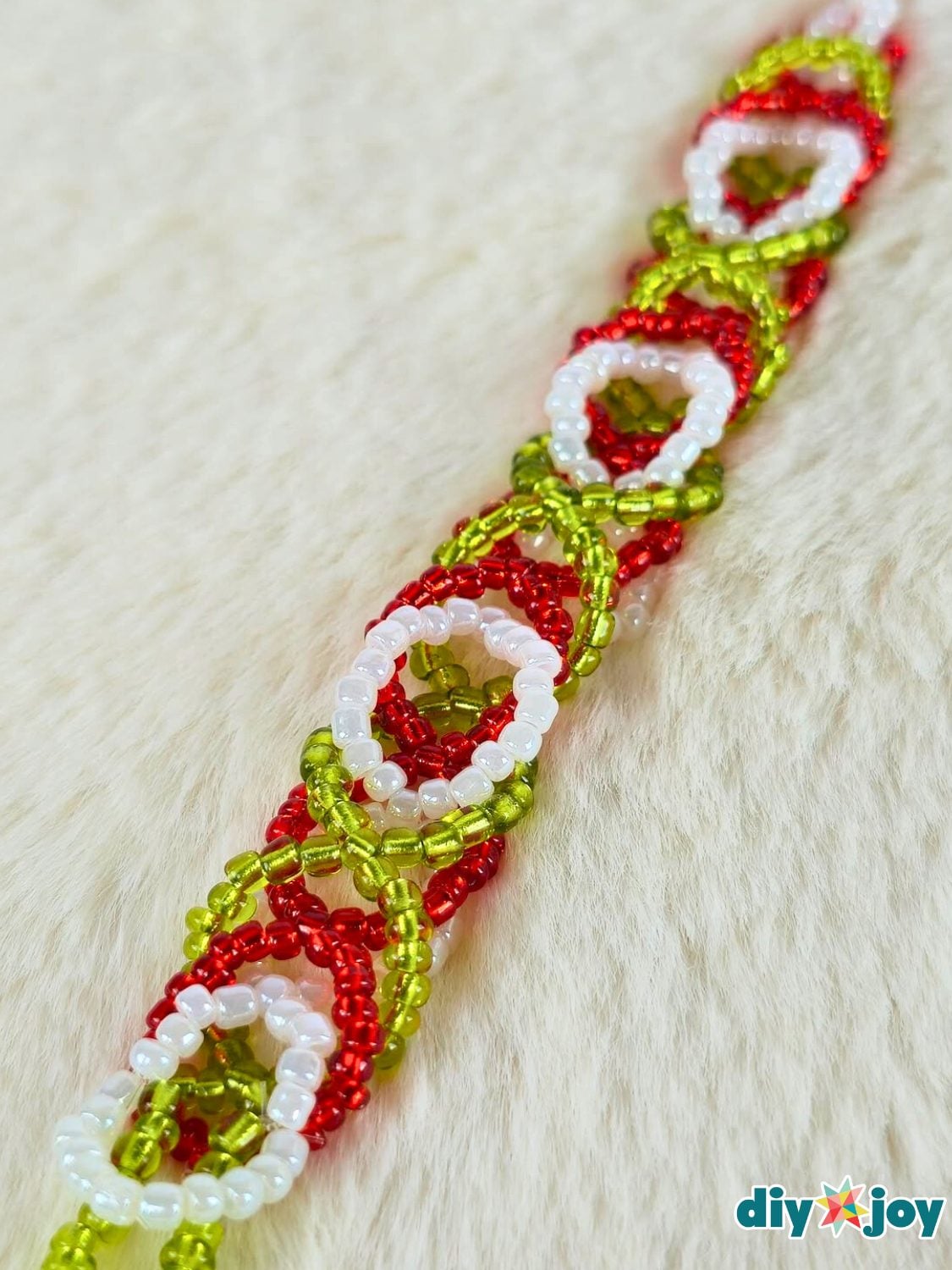 A woven seed bead bracelet in red, green and white with a small clasp lying on a white surface beside loose beads