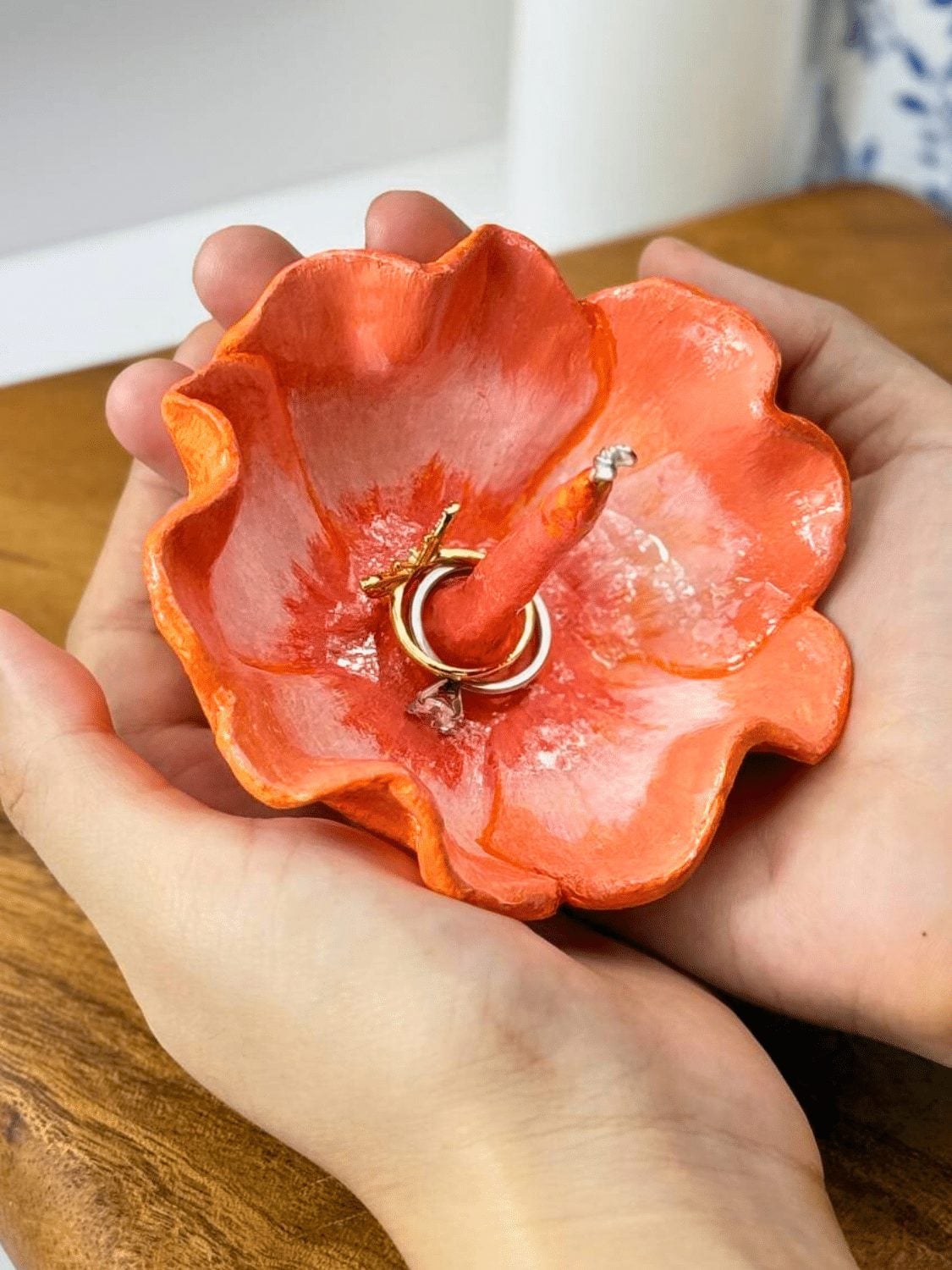 Two hands holding a coral painted air dry clay ring holder shaped like a flower with a center cone holding two gold rings