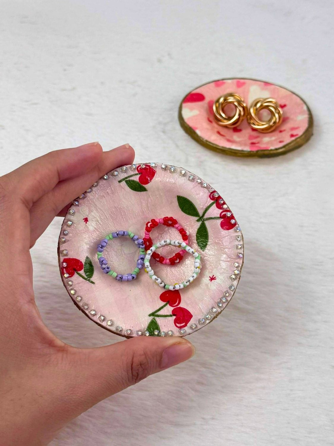 A hand holding a small clay ring dish decoupaged with cherry print paper and edged in rhinestones, a heart print dish behind