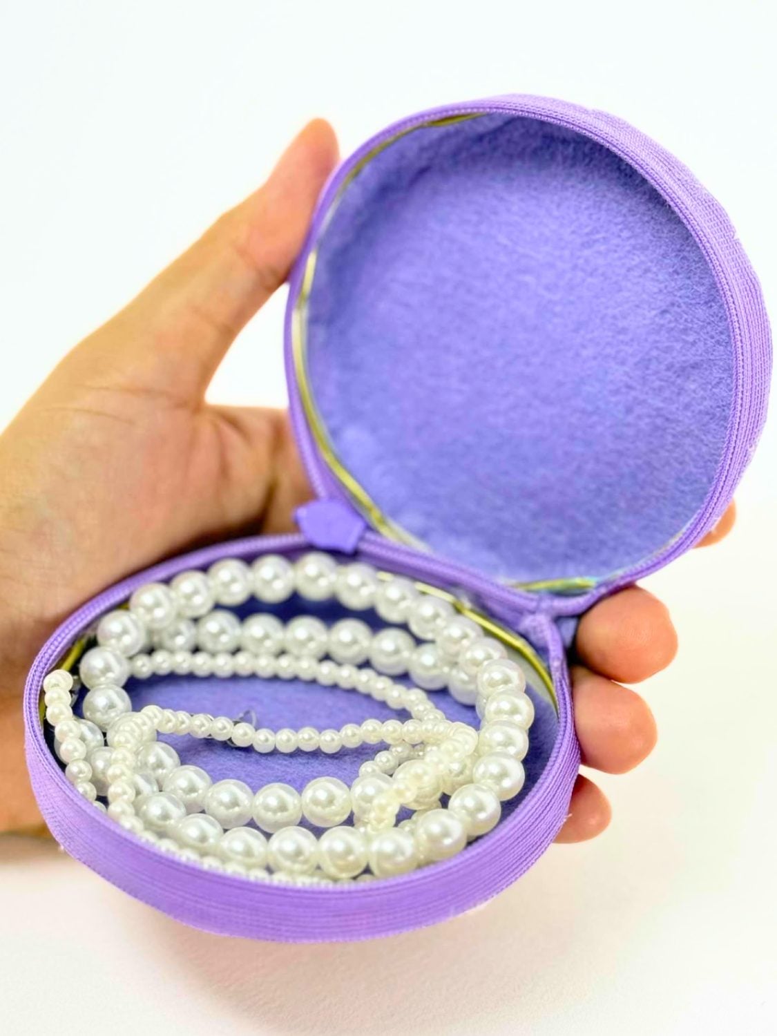 A hand holding an open lavender fabric jewelry purse made from two jar lids, lined with felt and holding a pearl necklace