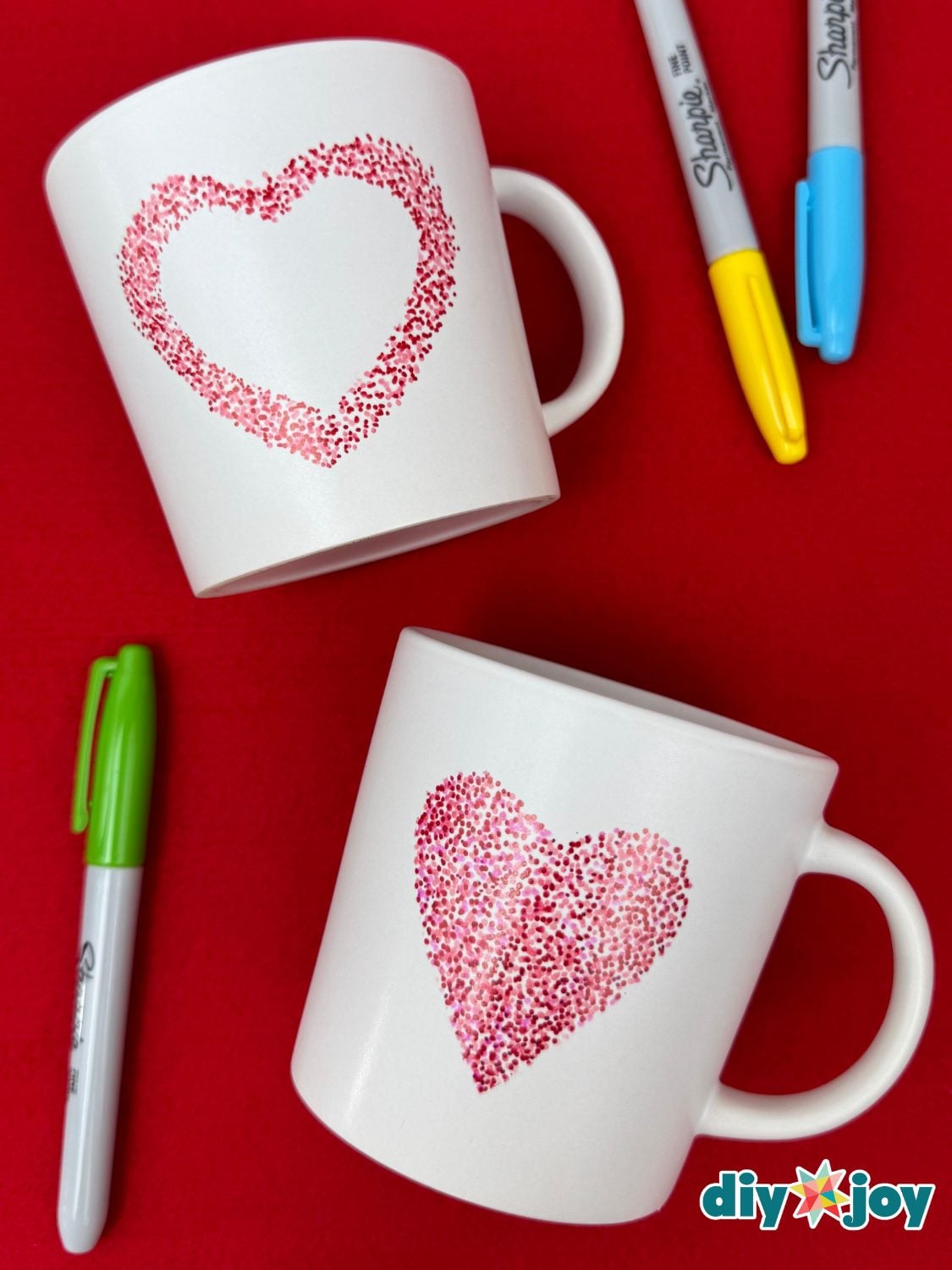 Two white mugs decorated with hearts made of tiny red and pink dots, one solid and one outlined, beside paint markers on red