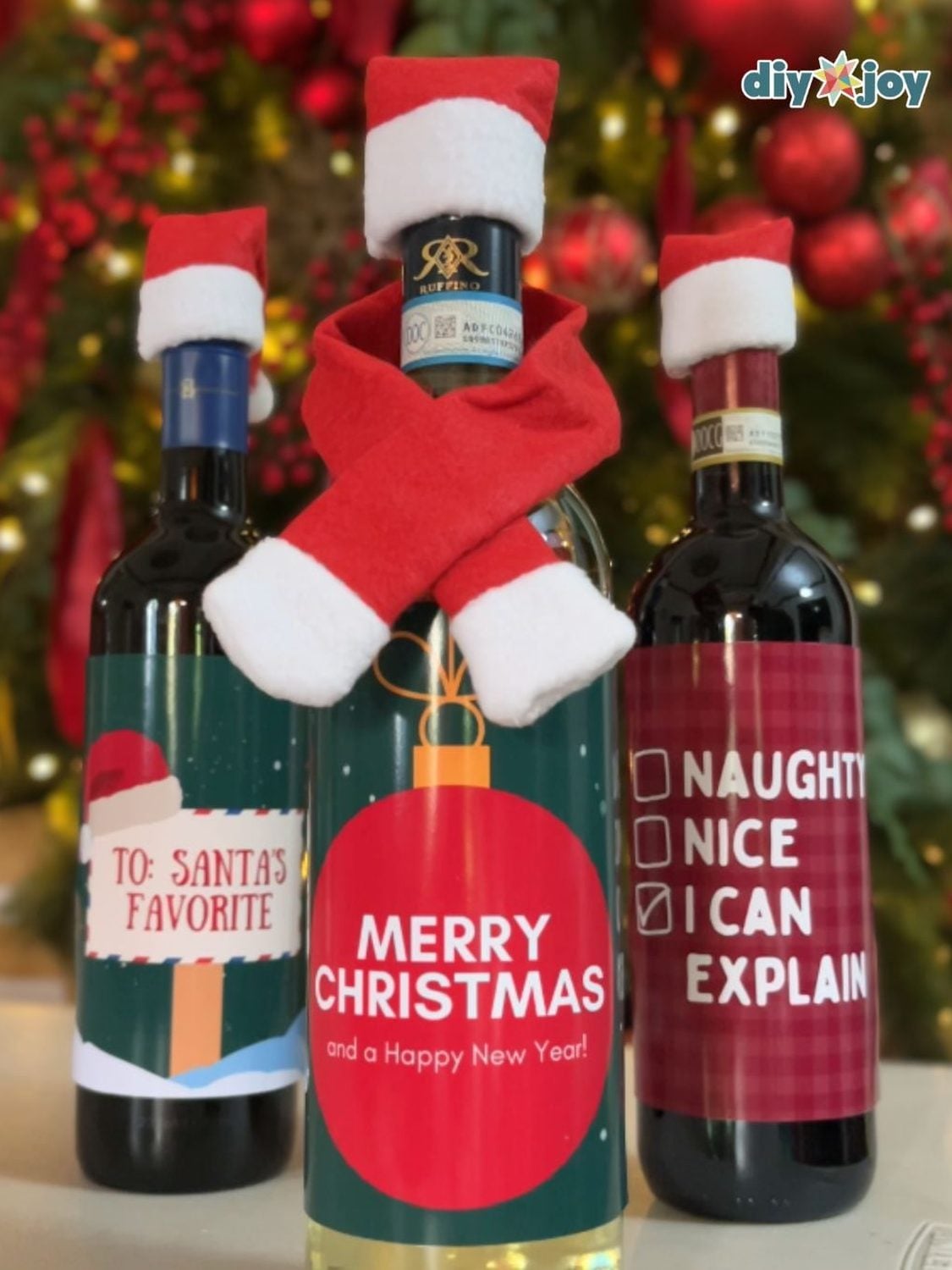 Three wine bottles in tiny felt Santa hats and a red scarf with printed labels reading Merry Christmas and I can explain