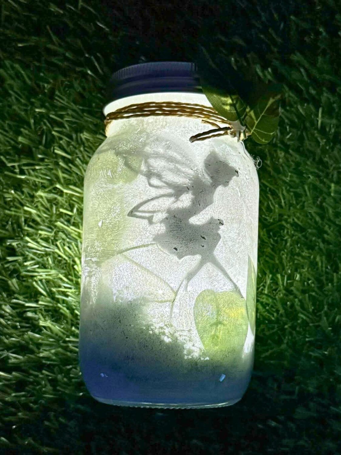 DIY Fairy Jar Lantern Night Light A frosted mason jar lantern glowing pale blue and white with a fairy silhouette inside, tied with twine and a leaf on grass
