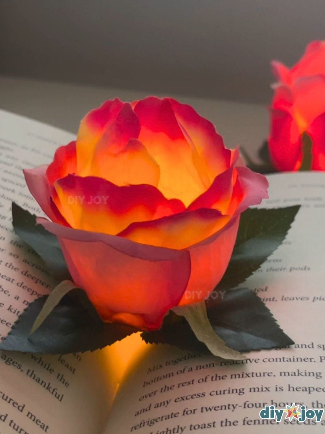 DIY Glowing Flower Tea Light Decor A red and gold faux rose glowing from a tea light inside its petals on an open book, with a second glowing rose behind