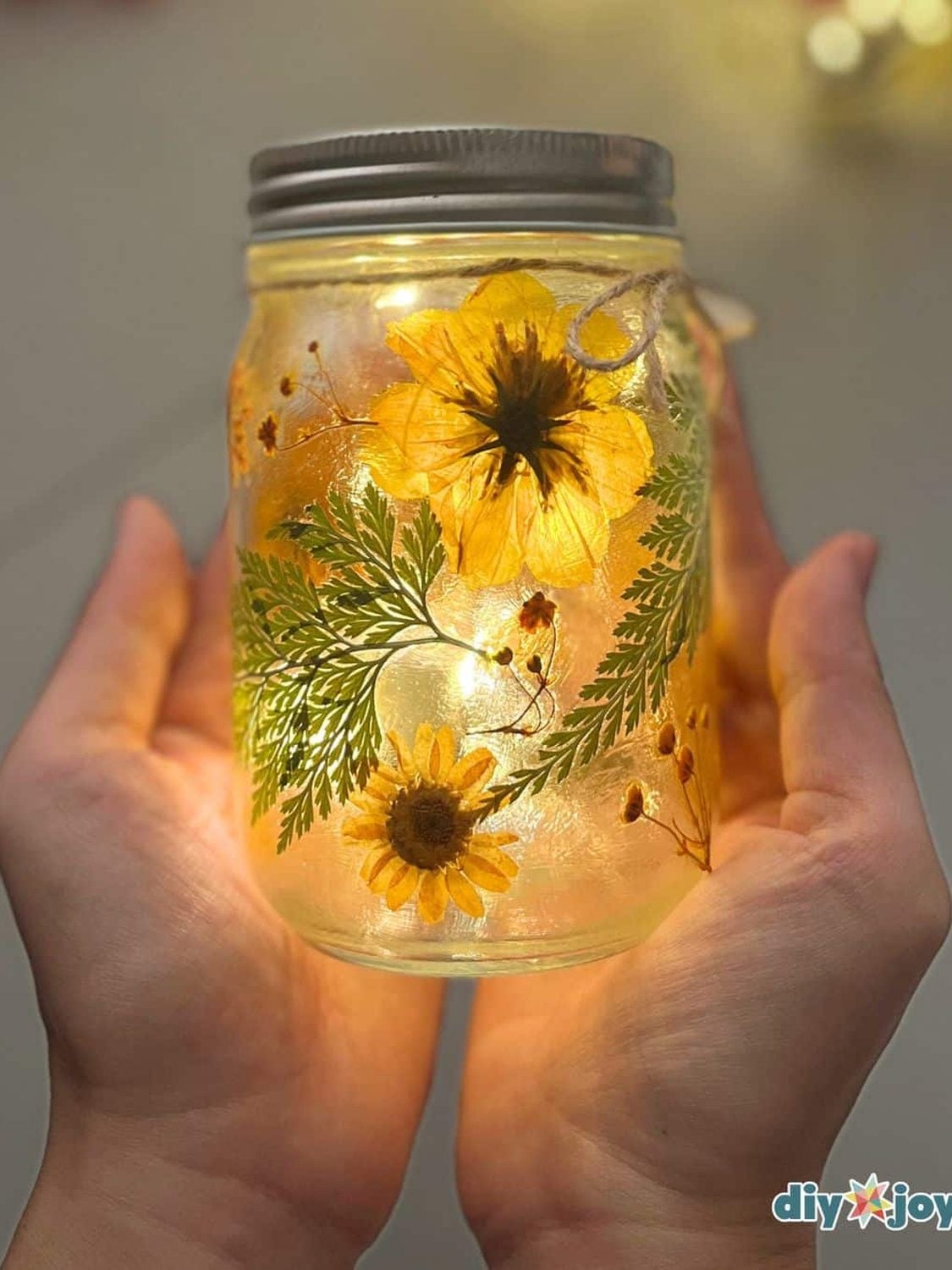 Pressed Flower Mason Jar Fairy Lights A glass mason jar covered in pressed flowers and leaves with fairy lights inside making the petals glow in a dark room