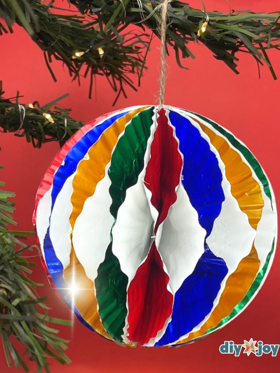 A striped ornament of folded metallic cupcake liners in red, blue, green and gold hanging by twine on a lit tree branch