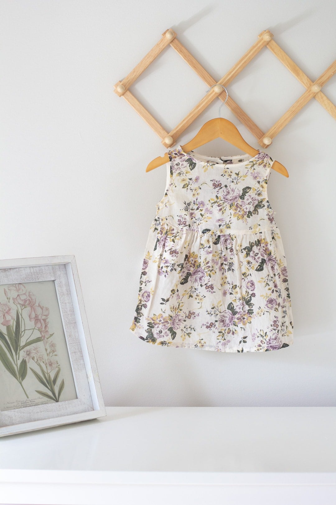 Small floral baby dress on a wooden hanger against a white wall, hanging beside a framed botanical print