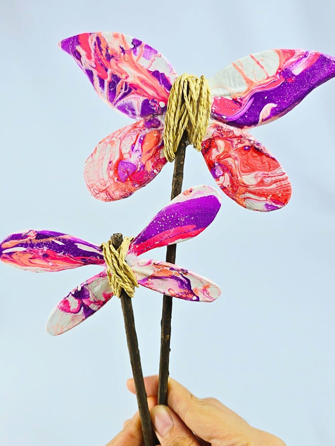 A marbled pink and purple clay butterfly and dragonfly on twig wands wrapped with twine, held up against a pale blue sky