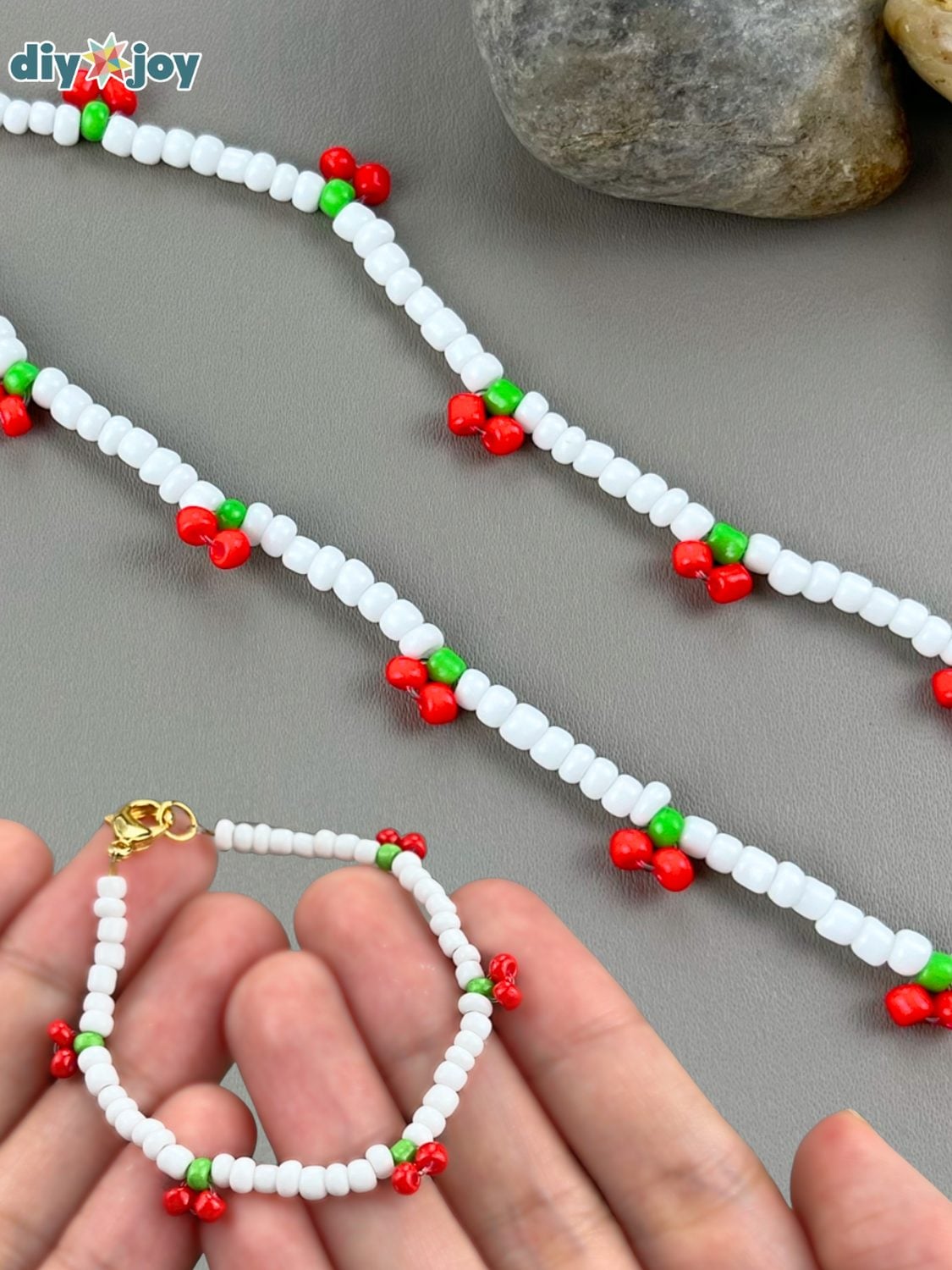 A white seed bead bracelet with tiny red and green cherry clusters and a gold clasp held in a hand beside matching necklaces