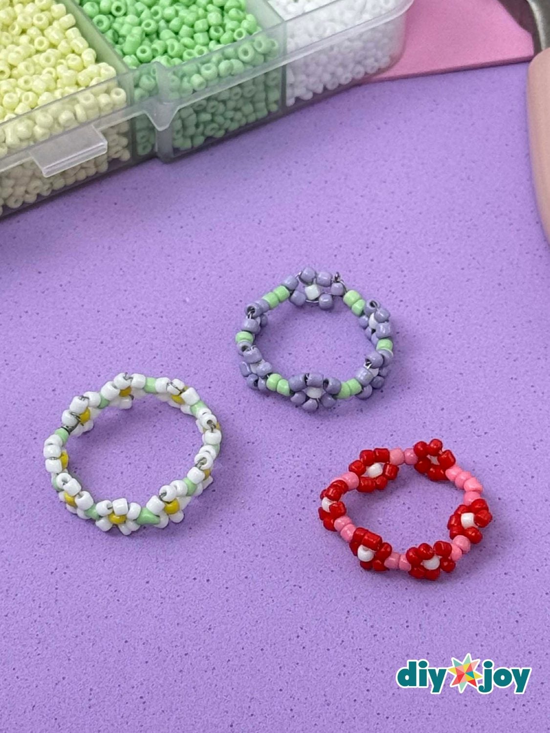 Three seed bead daisy rings in white and yellow, lavender and green and red and pink on a purple mat beside a bead box