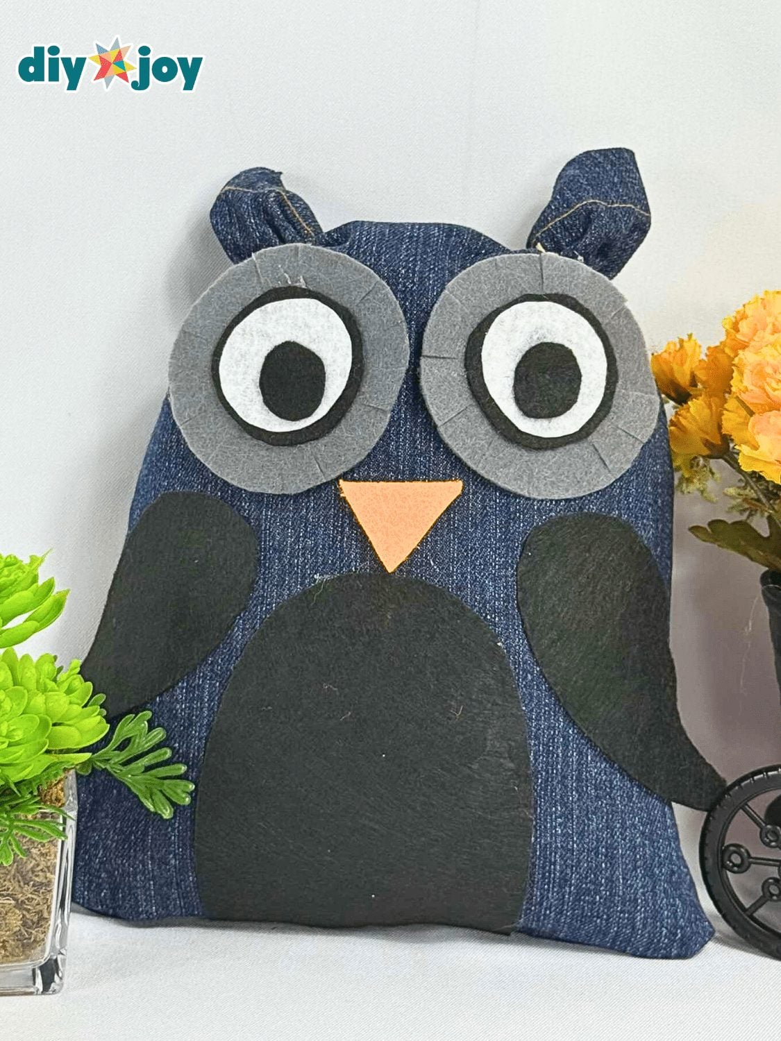 A stuffed owl cut from dark denim with layered gray, black and white felt eyes, an orange beak and dark felt wings on white