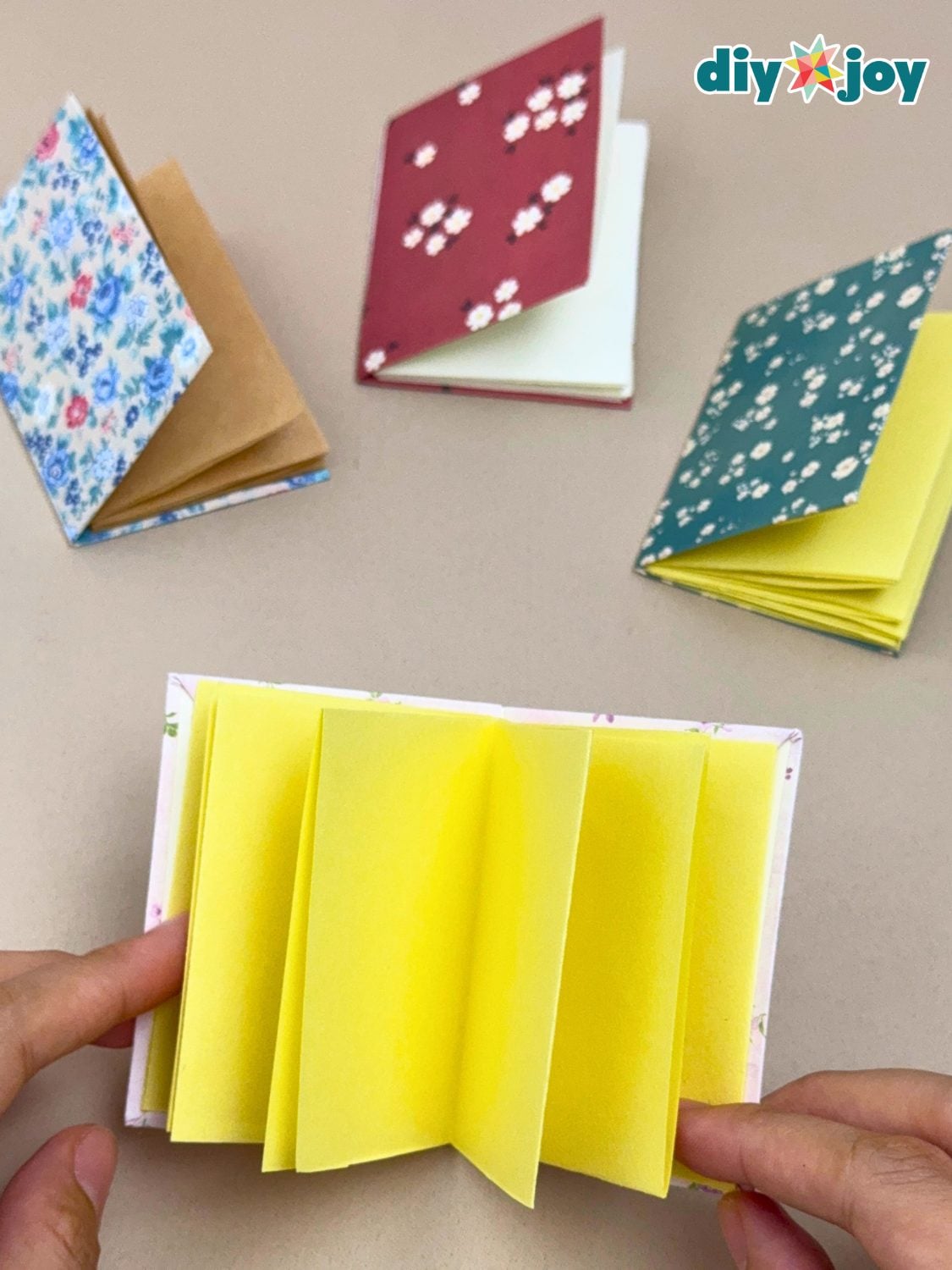 Four tiny handmade notebooks with floral scrapbook paper covers and folded yellow and kraft pages, one held open