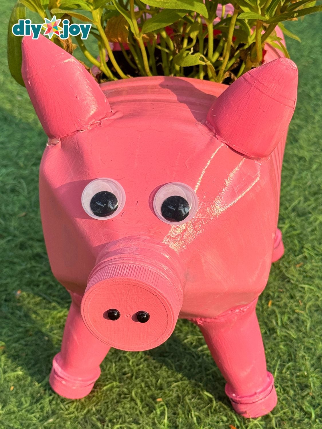 A pink pig planter made from a plastic jug with googly eyes, a bottle cap snout and a plant growing from its back on grass