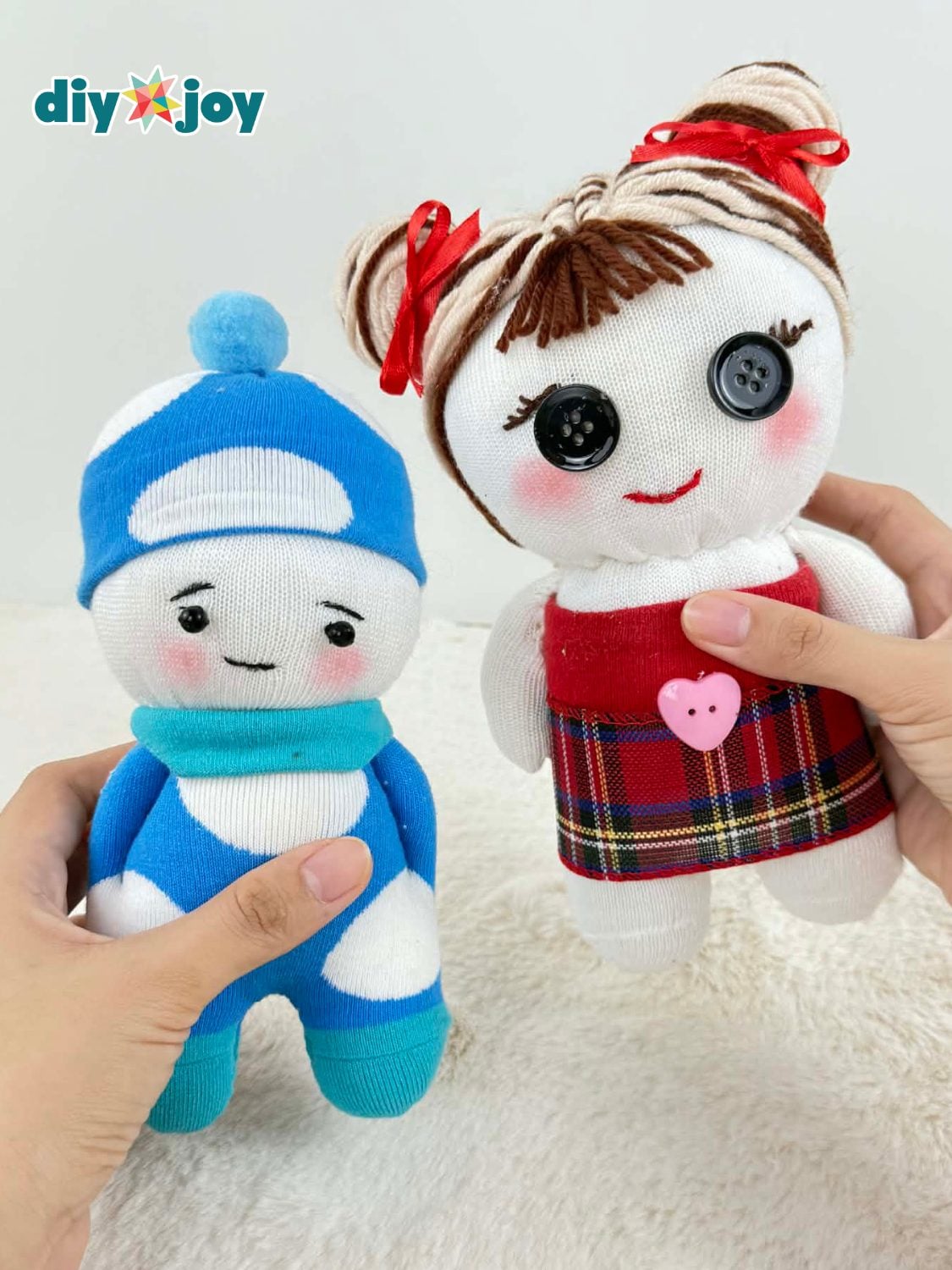 Two handmade sock dolls, one in a blue pom pom hat and one with yarn pigtails, red bows and a plaid skirt, held on white