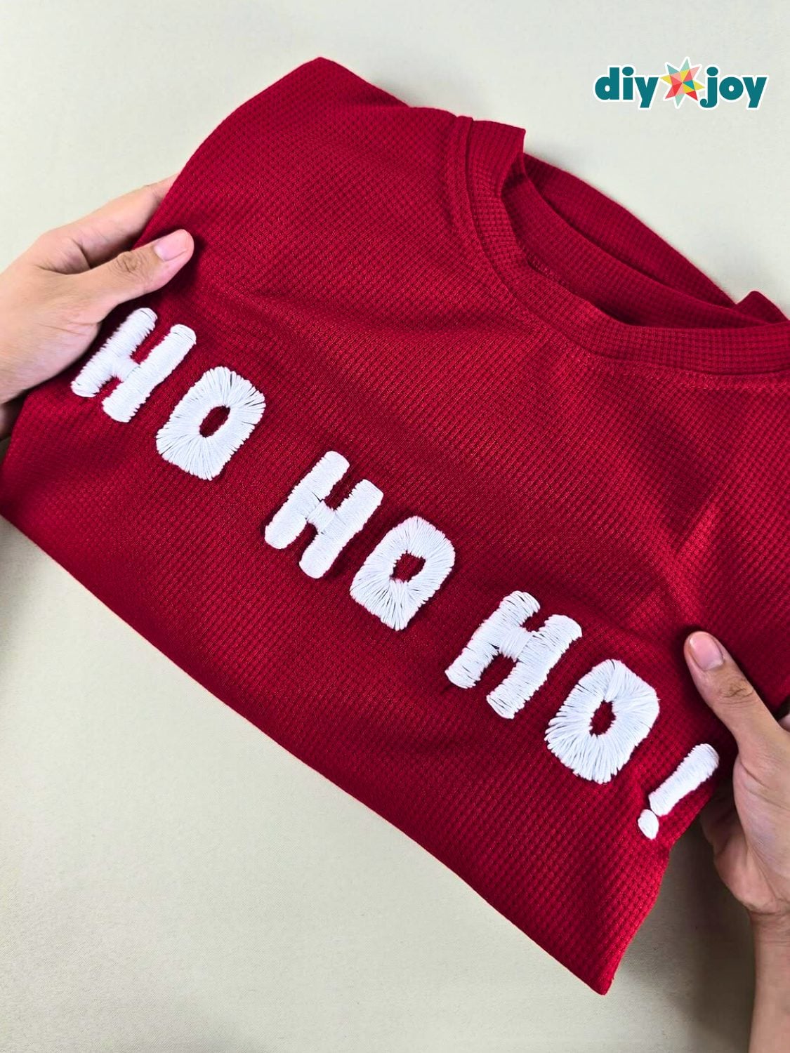 A folded red waffle knit sweater held in two hands with HO HO HO stitched across the chest in raised white cord letters