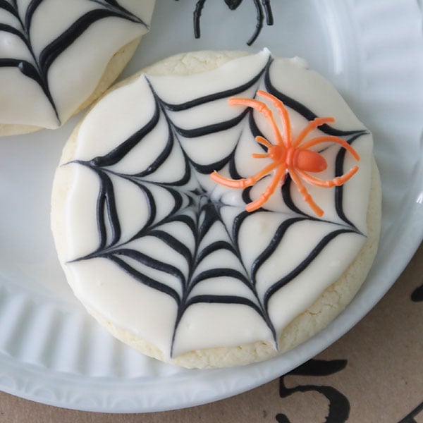 Easy Spiderweb Sugar Cookie Design Round sugar cookie with white frosting, a black piped web and a small spider decoration on top for Halloween