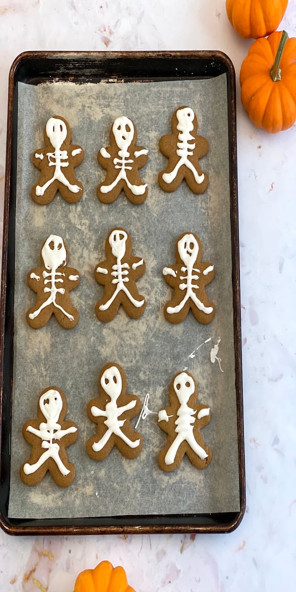 Skeleton Gingerbread Halloween Cookies Gingerbread people piped with white skeleton bones and faces beside small pumpkins on a baking sheet