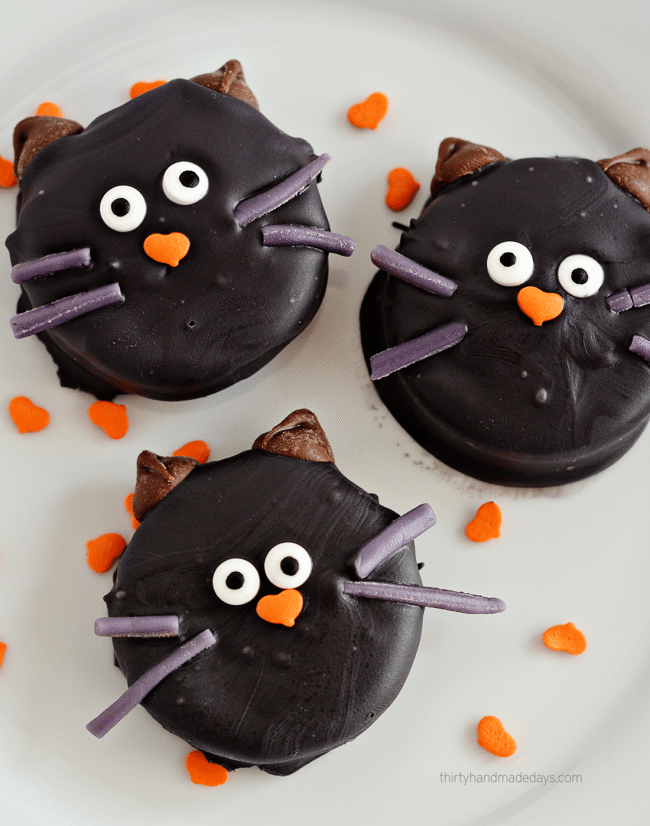 Black Cat Oreo Halloween Cookies Three black Oreo cat cookies with candy eyes, pointed ears and whiskers arranged on a white plate for Halloween