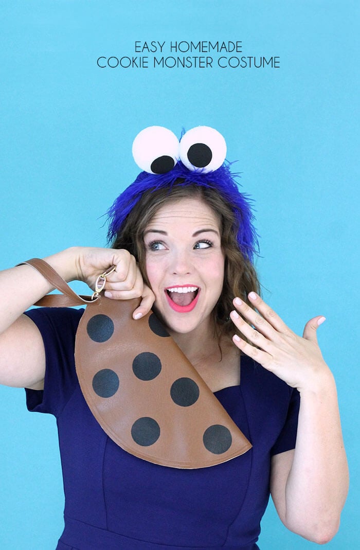 Homemade Cookie Monster Halloween Costume Adult wearing a blue Cookie Monster eye headband and holding a brown chocolate-chip cookie clutch against a blue backdrop