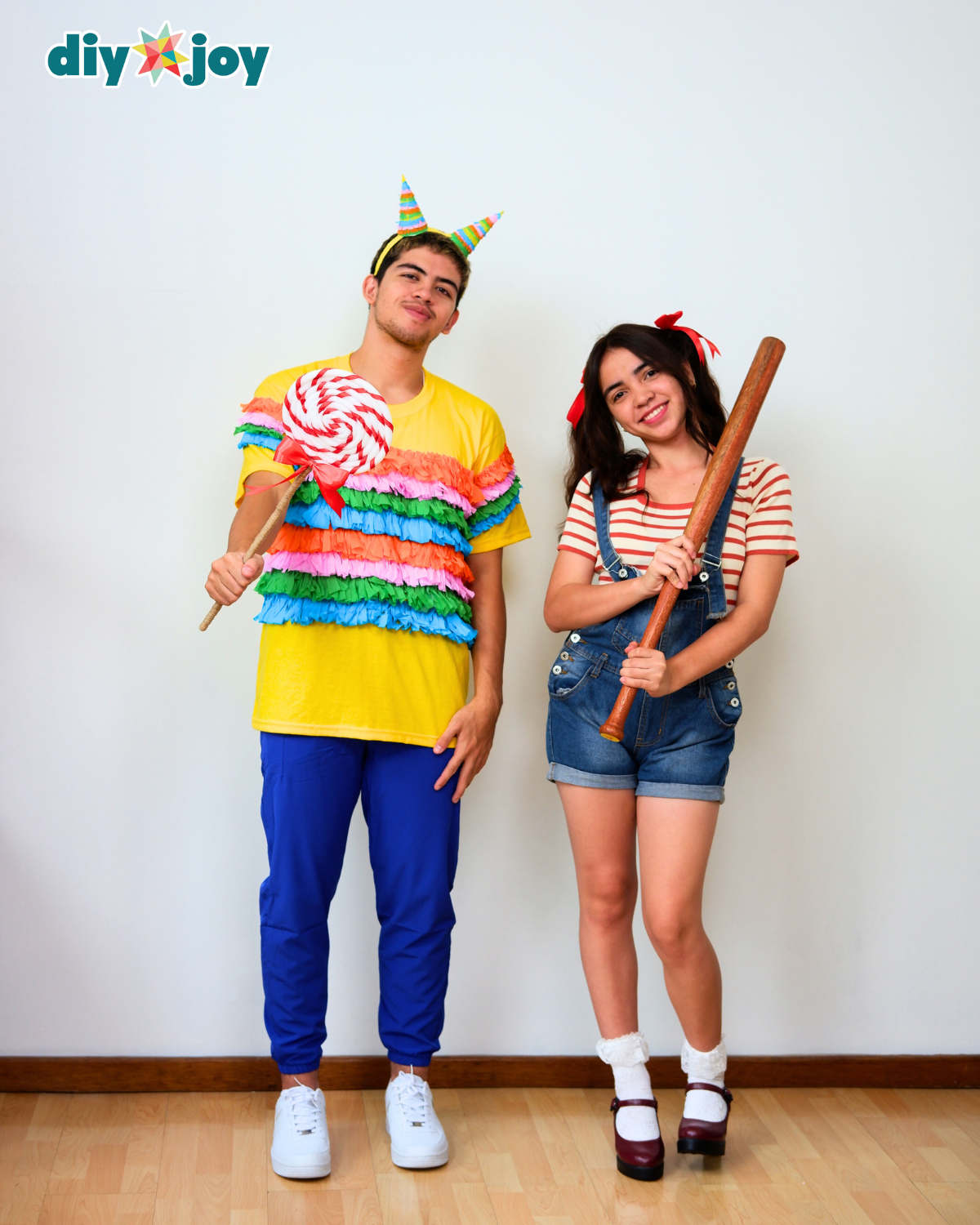 Piñata and Candy Halloween Costume Two adults wearing colorful piñata and candy costumes with striped fringe, candy prop and wooden stick against a white wall