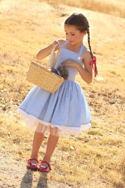 Dorothy Gingham Dress Halloween Costume Child wearing a blue gingham Dorothy dress with red shoes and woven basket outdoors with Girl Inspired source mark