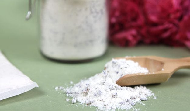 Wooden scoop of white lavender flecked milk bath soak spilled beside a filled glass jar, an easy homemade gift in a jar