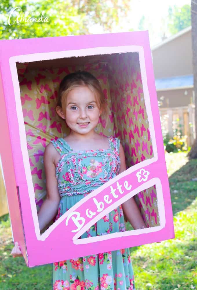 DIY Cardboard Doll Box Costume Child wearing a pink doll box costume made from cardboard with painted trim and patterned paper lining