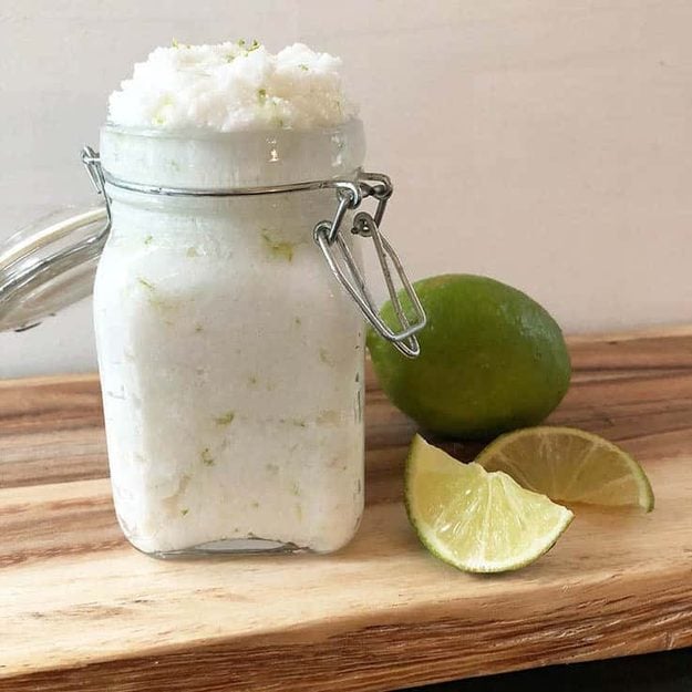 Coconut lime body scrub heaped in a swing top glass jar on a wood board with a whole lime and two lime wedges beside it