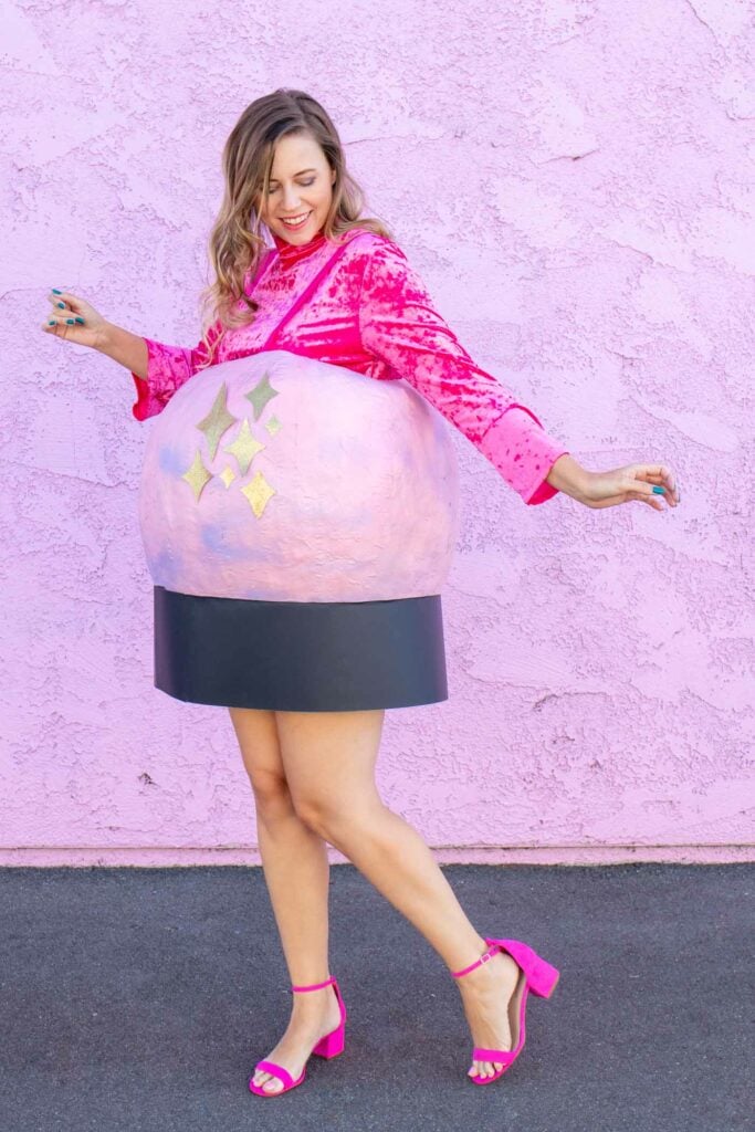 DIY Crystal Ball Halloween Costume Adult wearing a pink crystal ball costume with gold stars and black base over a pink velvet top near a pink wall
