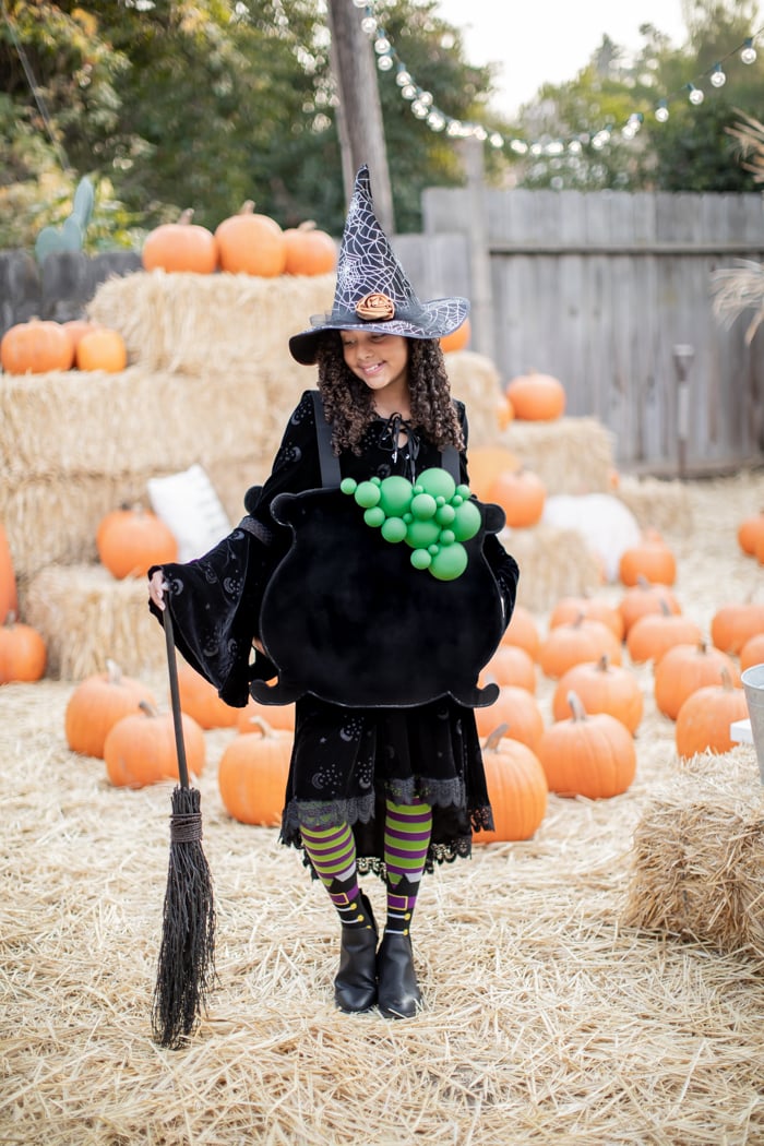 DIY Cardboard Cauldron Costume Child dressed as a witch wearing a black cauldron costume with green bubbles, orange flames and a broom outdoors