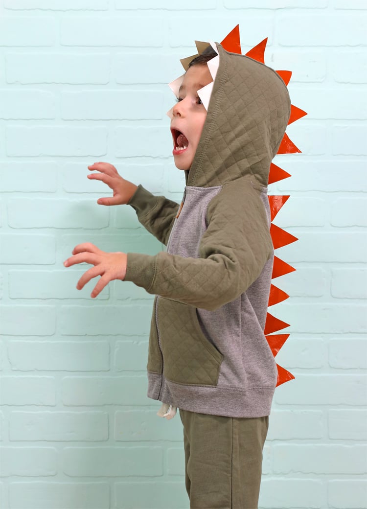 Easy No-Sew Dinosaur Costume Idea Child wearing a hooded orange dinosaur costume with white taped teeth and triangular spikes running down the back