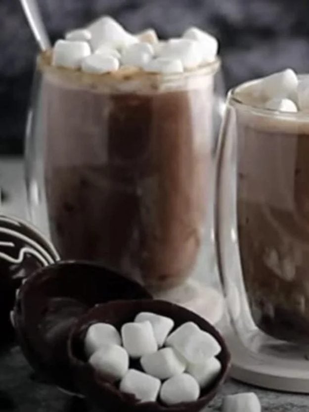 Two glass mugs of hot chocolate topped with mini marshmallows beside a cracked open chocolate hot chocolate bomb full of marshmallows