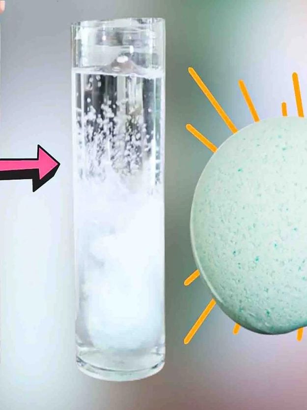 Bath bomb fizzing in a tall glass of water beside a pale teal speckled bath bomb, from a homemade bath bomb tutorial
