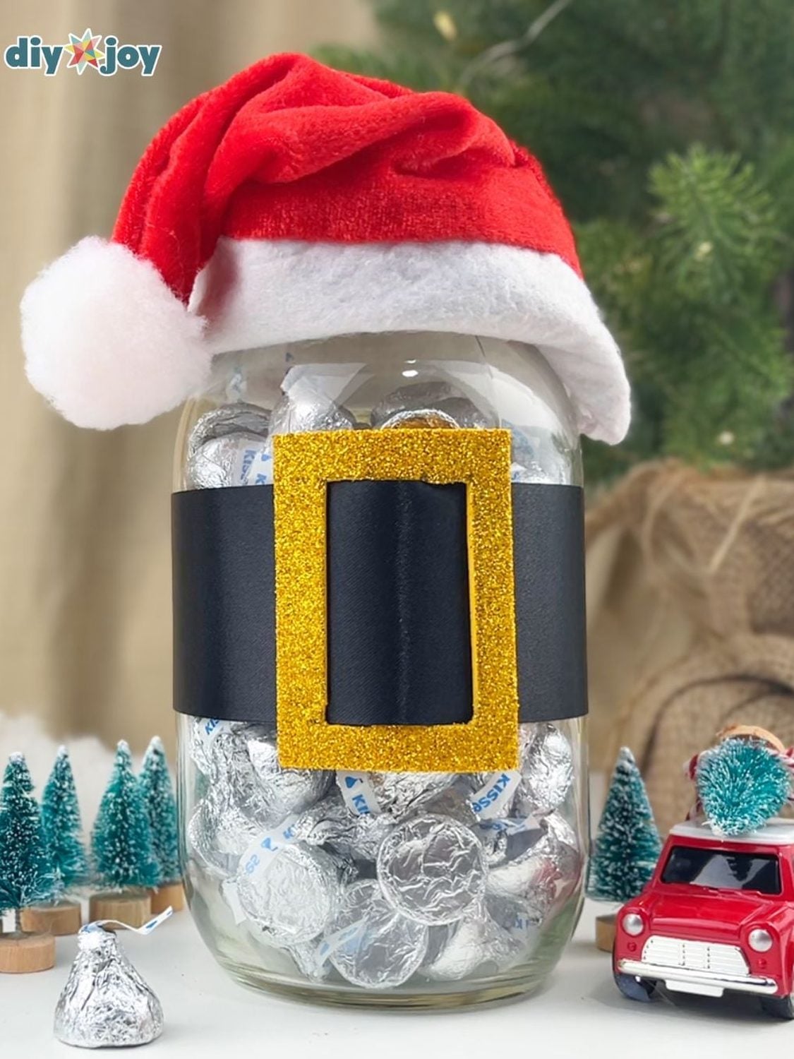 Mason jar dressed as Santa with a red hat lid, black ribbon belt and gold buckle, filled with wrapped chocolates