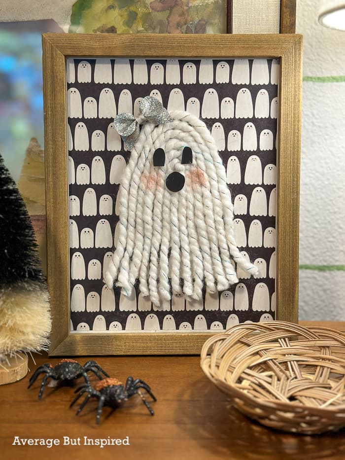Framed yarn ghost art with a macrame-look arch, black cardstock face, optional bow and rosy cheeks on patterned paper