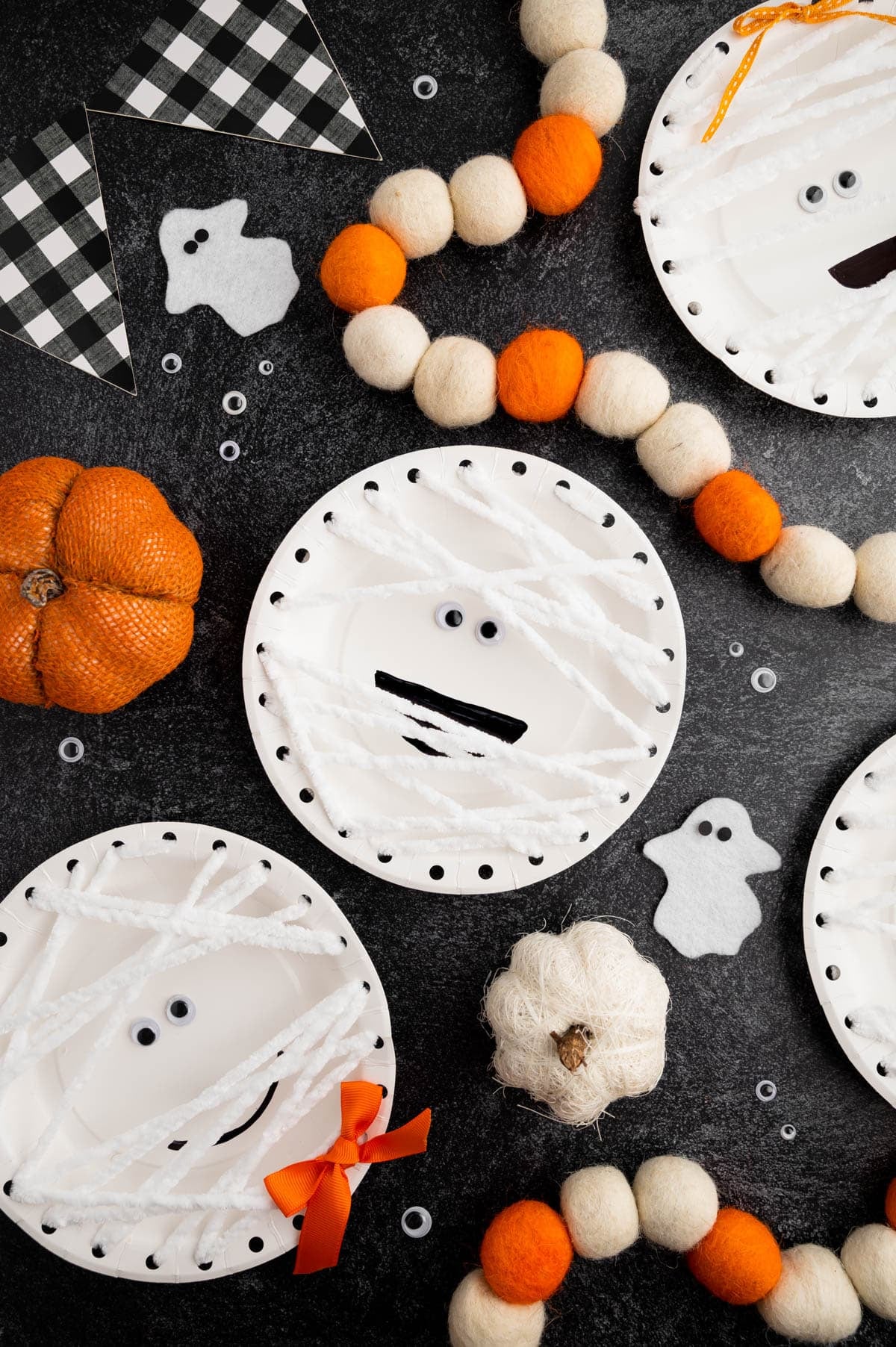 White paper-plate mummy crafts laced with white yarn, googly eyes and black mouths among orange Halloween decorations