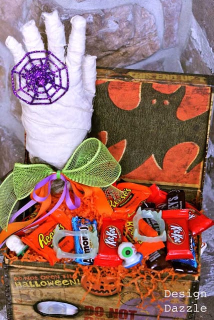 White wrapped mummy hand stands in a decorated candy box with purple web, ribbon and a Design Dazzle watermark