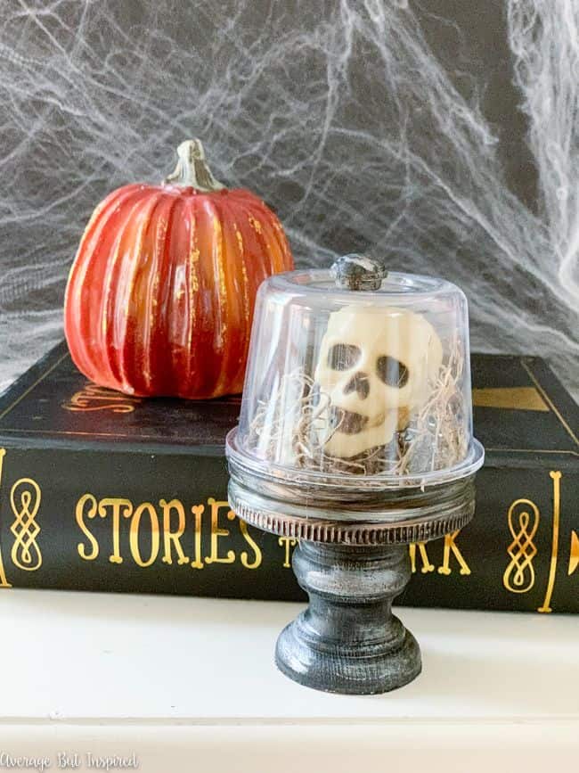 Miniature skull and Spanish moss sit under an inverted glass jar on a black and silver painted pedestal