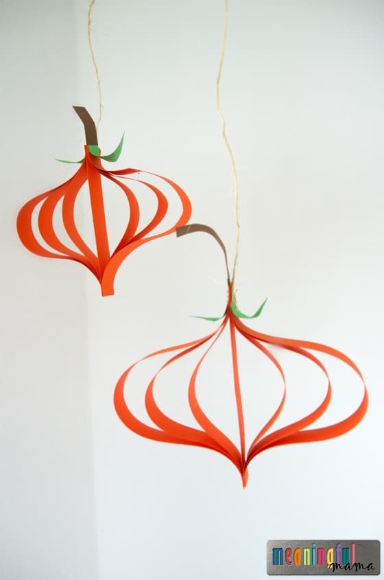 Two orange paper strip pumpkins hang against a light wall with brown stems and curled green paper vines