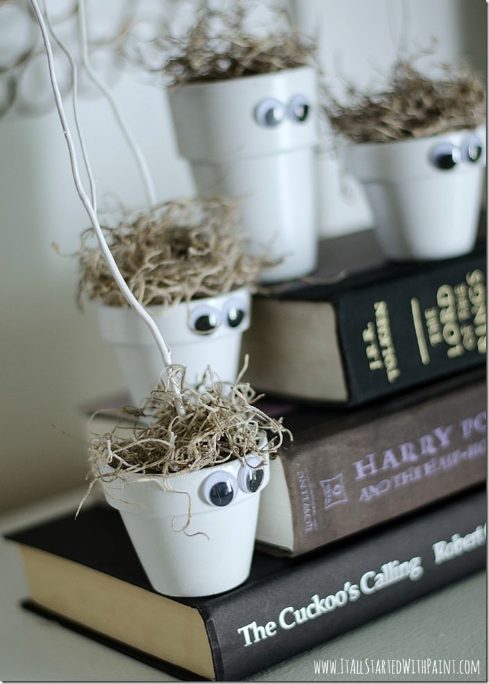 Four white terracotta monster pots with googly eyes, moss hair and tall white sticks sit on stacked books