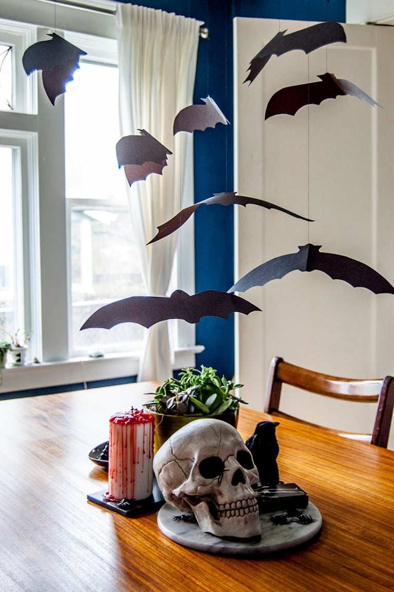 Multiple black paper bats hang from fine thread above a dining table arranged with Halloween decorations